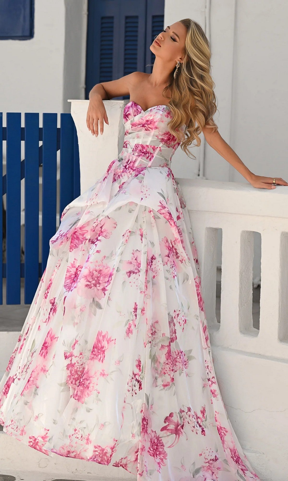 A woman poses outdoors in bright daylight, leaning against a white wall with blue accents, wearing the Portia and Scarlett PS26083 long formal dress in white/pink.